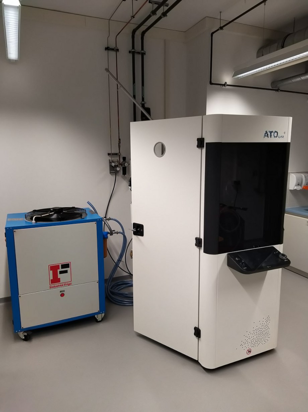 Metal powder atomization system ATO Lab Plus consisting of a white rectangular main unit with a black front door and a blue cubic auxiliary unit with cables in a laboratory room.