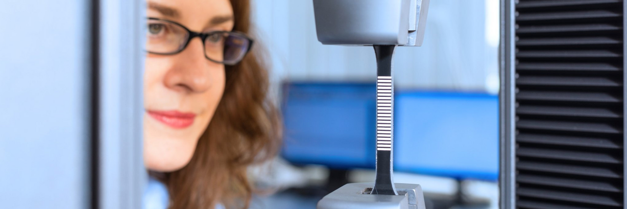Scientists wearing glasses operating a tensile testing machine with a metal test specimen clamped in a materials testing laboratory.