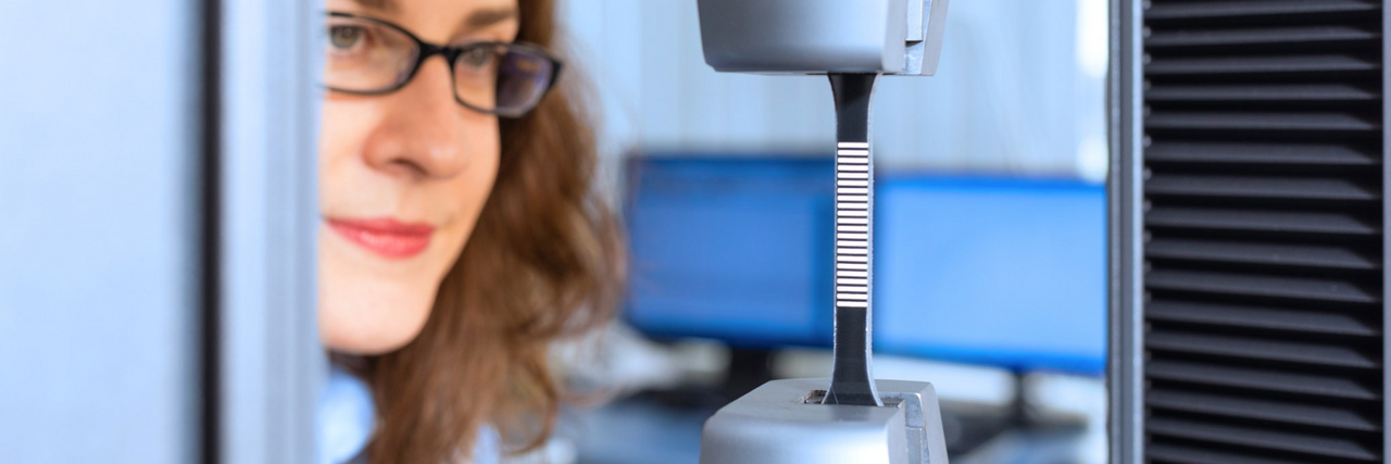 Wissenschaftlerin mit Brille bedient eine Zugprüfmaschine mit eingespanntem Metallprüfkörper in einem Labor für Werkstoffprüfung.