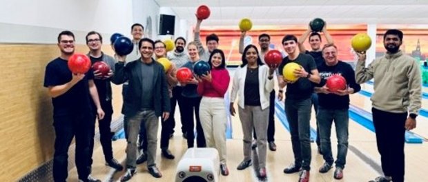 Members of the "Molecules To Materials"-Researchgroup standing in a bowling alley holding bowling balls with some raising them above their heads.