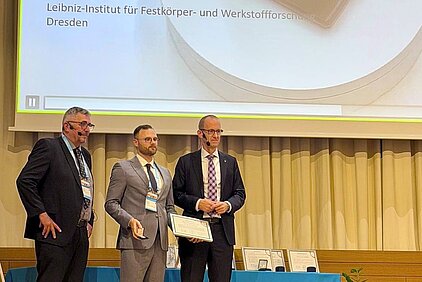 Three men in suits stand on a stage in front of a curtain, one holding a certificate, with a screen displaying text in the background.