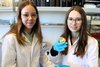 Zwei Mädchen mit weißen Laborkitteln und blauen Gummihandschuhen halten einen präparierten Probenhalter ins Bild.