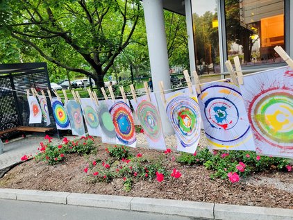 Multiple colorful target sheets hanging on a clothesline outdoors above a flower bed with red flowers. Pictures on clothesline showcase the creativity from this SpinBike Station by kids at the IMC at IFW Dresden.