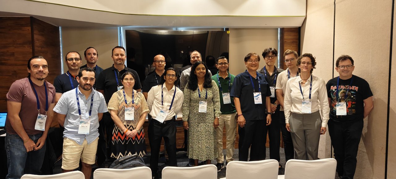 A group of people are posing for a group photo in a conference room. They are wearing name badges on lanyards around their necks.