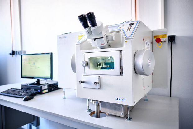 A benchtop unit for the powder-bed laser sintering process, fitted with a microscope attachment and a viewing window, positioned on a table next to a computer screen in a well-lit room.