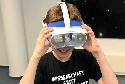 Junge mit Virtual-Reality-Brille und T-Shirt mit der Aufschrift 'WISSENSCHAFT STATT NACHT' lächelt.