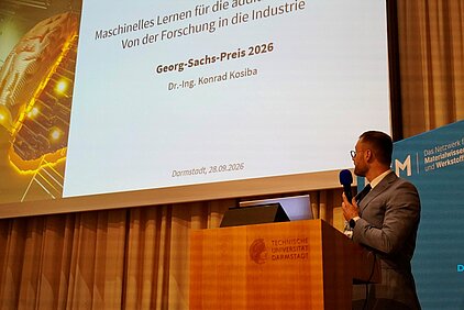 Dr. Konrad Kosiba in a suit holding a microphone stands at a podium in front of a screen projecting a title about machine learning and the Georg Sachs Prize 2026.