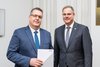 Zwei Herren, gekleidet im Anzug, stehen nebeneinander. Der Herr links zeigt eine Urkundenmappe in die Kamera. 