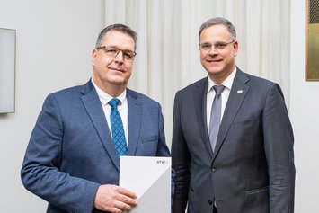 Zwei Herren, gekleidet im Anzug, stehen nebeneinander. Der Herr links zeigt eine Urkundenmappe in die Kamera. 