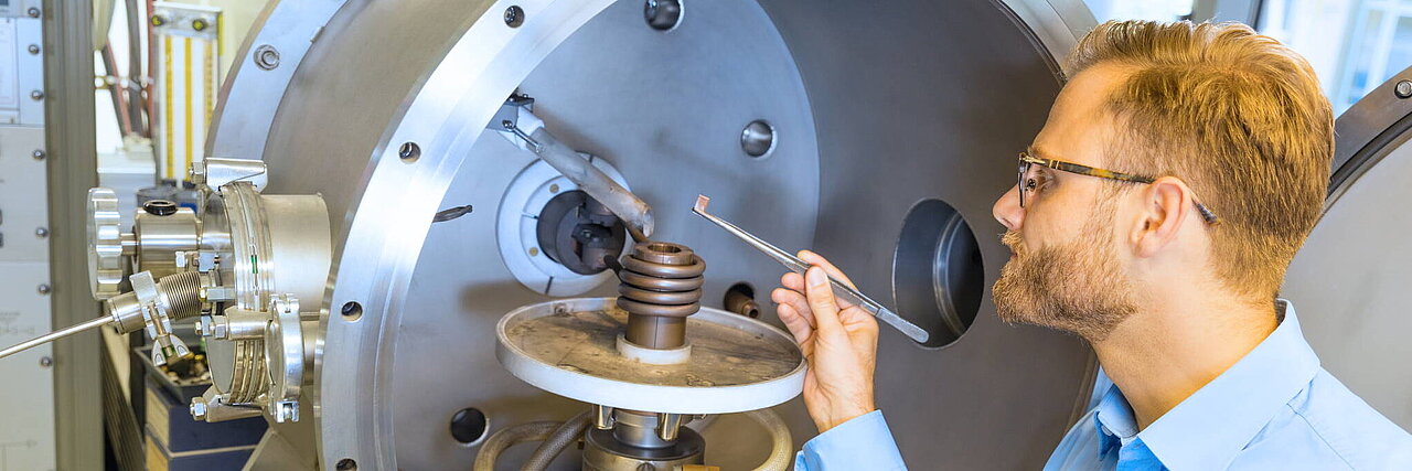 [Translate to Deutsch:] Man in a blue shirt stands in front of a large metal chamber and holds a tool to a spiral device inside.