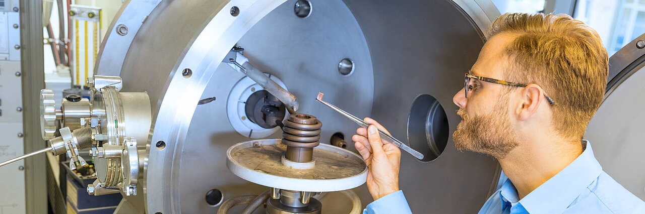 Man in a blue shirt stands in front of a large metal chamber and holds a tool to a spiral device inside.