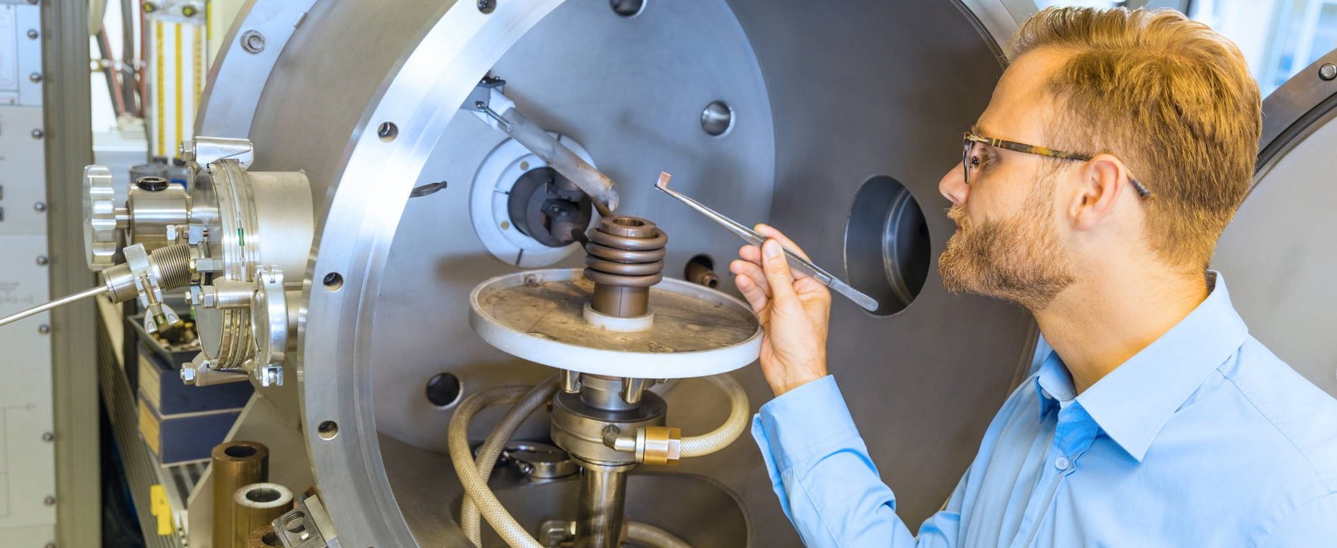 Man in a blue shirt stands in front of a large metal chamber and holds a tool to a spiral device inside.