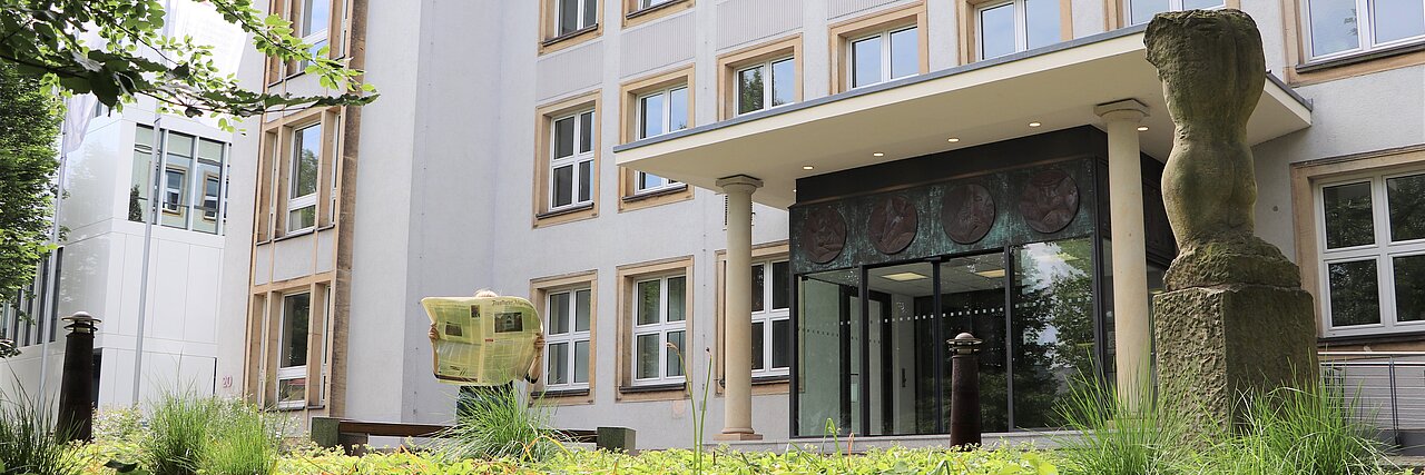 Eine Person mit einer großen Zeitung sitzt vor dem Hauptportal des IFW Dresden. Am unteren Bildrand sind die Büsche des Vorplatzes sattgrün zu sehen.