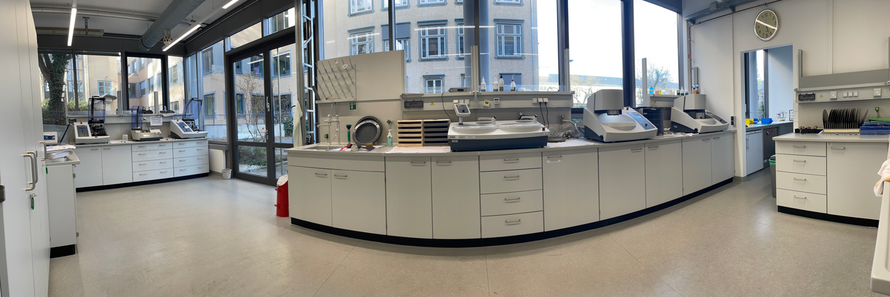 Laborgerät zur metallographischen Präparation auf einer langen Laborarbeitsfläche vor Fenstern mit Blick nach draußen.