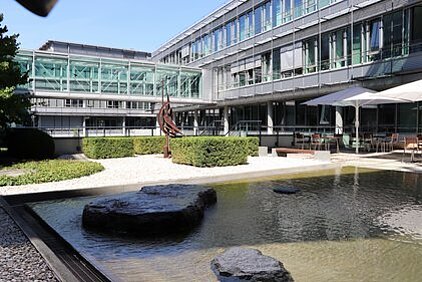 Courtyard of a modern building complex of the IFW with a water pool containing large stones, a sculpture, and seating areas with umbrellas.