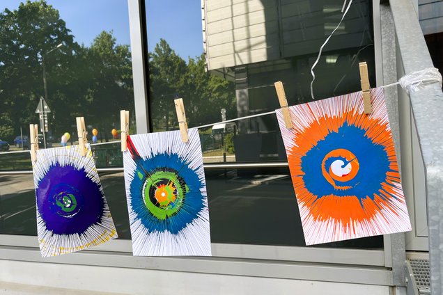 Three abstract spiral paintings with radial lines in blue, green, yellow, and orange colors hanging on a string with clothespins outside a building window at SpinBike station at IFW.