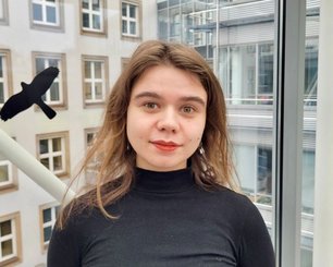 Pauline Becker with shoulder-length wavy hair wearing a black turtleneck sweater standing in front of a building with large windows.