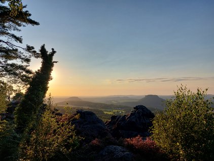 Landscape view scenic 5-kilometre hike to the Bergwirtschaft Papststein at sunset with rocky foreground, trees on the sides, and distant hills under a clear sky.