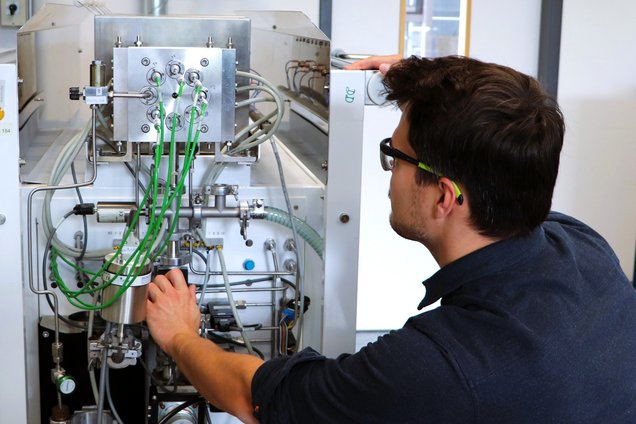 A young man standing with his back to a stainless steel unit fitted with various green hoses and connectors.