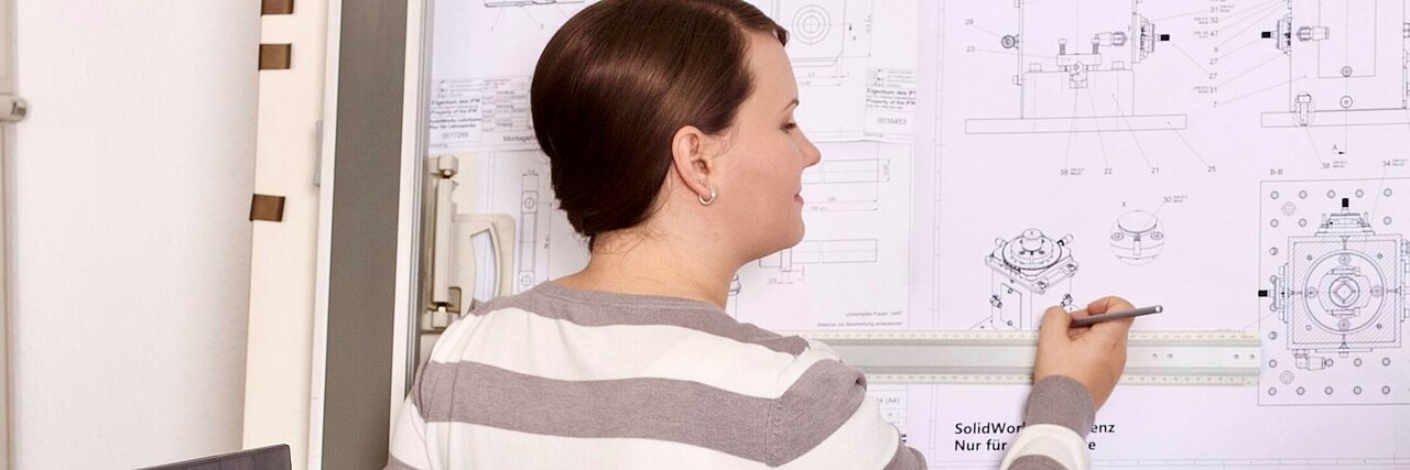 Produktdesignerin mit Zopf und gestreiftem Pullover steht vor einer Wand mit technischen Zeichnungen und hält einen Stift in der rechten Hand.