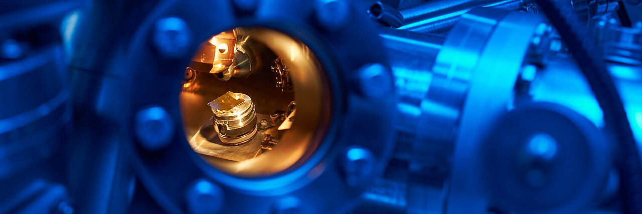 View through a circular opening onto a complex apparatus with metallic components and an illuminated chamber inside.