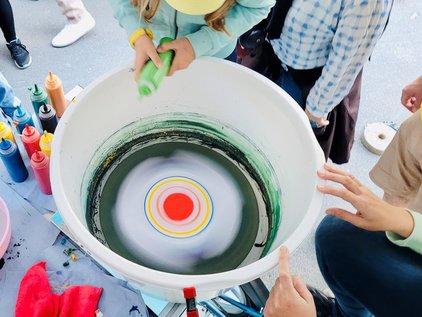 Top view of a bucket with a red-yellow-green color wheel surrounded by several hands and paint bottles. It shows the setup at work during the SpinkBike Sessions at LNDWDD at IMC.