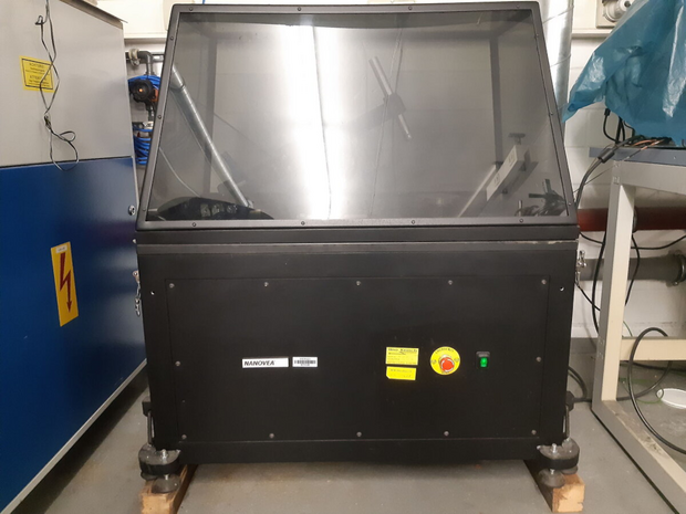 Black laboratory device with transparent lid, yellow emergency stop button, and green button on the front, standing on gray floor.