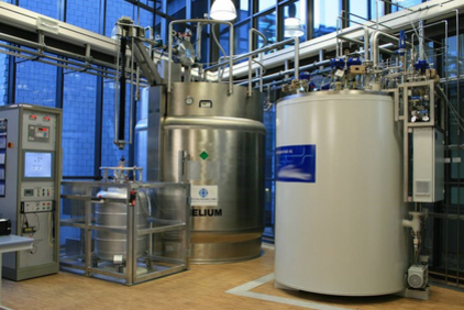 Large metallic laboratory equipment and containers in a technical room with pipes and control units, including a container labeled 'HELIUM'.