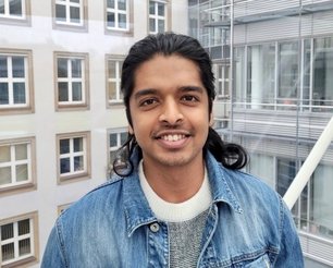 Bijul Mankara Sureshbabu with dark tied-back hair wearing denim jacket and sweater in front of modern building with large windows.