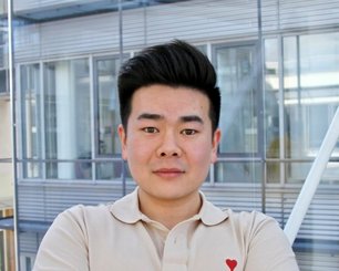 Xuan Qi with dark hair wearing a light beige polo shirt with a small red heart on the chest standing in front of a modern glass building.