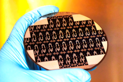 Hand wearing a blue glove holding a round silicon wafer with multiple small rectangular black microchip structures.