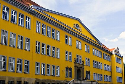 BSZ für Wirtschaft “Prof.-Dr.-Zeigner” in Dresden is a large yellow school building with many windows and a gable with a clock under a blue sky.