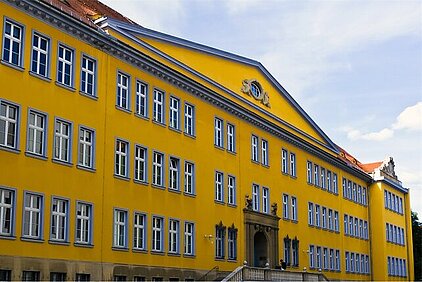 Das BSZ für Wirtschaft "Prof.-Dr.-Zeigner" in Dresden. BSZ für Wirtschaft "Prof.-Dr.-Zeigner" in Dresden ist ein großes, gelbes Schulgebäude mit vielen Fenstern und einem Giebel mit Uhr unter blauem Himmel.
