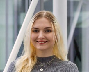 Julia Prodan with long blonde hair wearing a gray sweater and a necklace with a small pendant in front of a modern glass building.