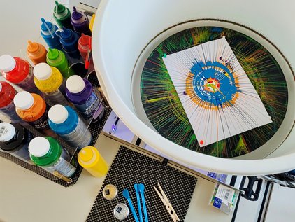 Table with multiple bottles of colorful paint, a white round frame with stretched threads and a paper with a circular pattern in the center, along with various tools and materials for thread art. Setup for SpinBike station at LNdWDD by IMC at IFW.