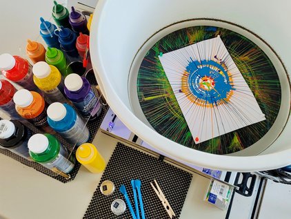 Table with multiple bottles of colorful paint, a white round frame with stretched threads and a paper with a circular pattern in the center, along with various tools and materials for thread art. Setup for SpinBike station at LNdWDD by IMC at IFW.