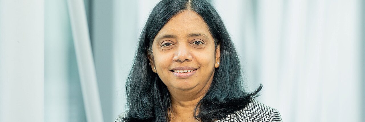 Porträt einer Frau, Prof. Dr. Anjana Devi, mit schulterlangen schwarzen Haaren und kariertem Blazer vor hellem, unscharfem Hintergrund.