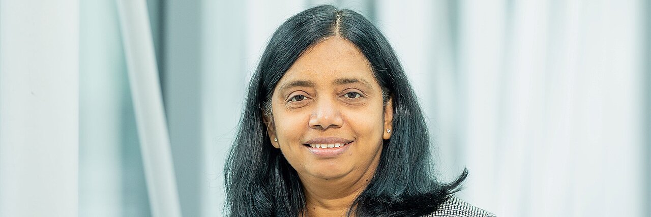 Portrait of a woman, Prof. Dr. Anjana Devi, with shoulder-length black hair wearing a checkered blazer against a light blurred background