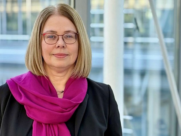 Head of office Brit Präßler-Wüstling with blond shoulder length hair and a purple scarf in front of bright window.
