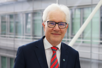 Portrait of theoretical physicist Prof. Vladimir Fomin at IFW Dresden in front of a modern glass-and-steel interior. He is wearing a dark suit, a white dress shirt, and a red striped tie.