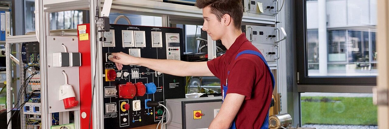 Young electrician in red T-shirt and blue overalls operating control panel in workshop with windows in the background.