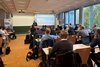 Menschen sitzen in einem Seminarraum. Vorn steht ein Dozent und trägt eine Präsentation vor.