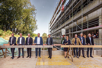 Group photo at the topping-out ceremony for the joint research building of IFW Dresden and TU Dresden: nine representatives from science, politics and construction stand with hammers in front of the ceremonial timber beam.