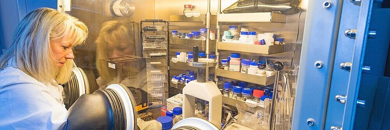 Technician in white lab coat working at a sealed glovebox with gloves and various containers in the background.