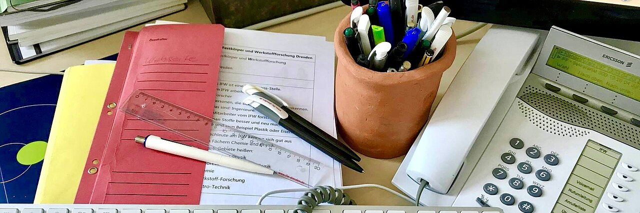 Schreibtisch eines Büroarbeiters mit Tastatur, Telefon, Stiftebecher, Aktenordnern und Dokumenten.