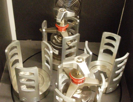 A Retsch planetary ball mill with its cover open, showing three cylindrical stainless steel grinding jars arranged on a circular platform. Each jar is wrapped in a slotted metal cage with rectangular cutouts. The components are silver-gray against a dark chamber interior.