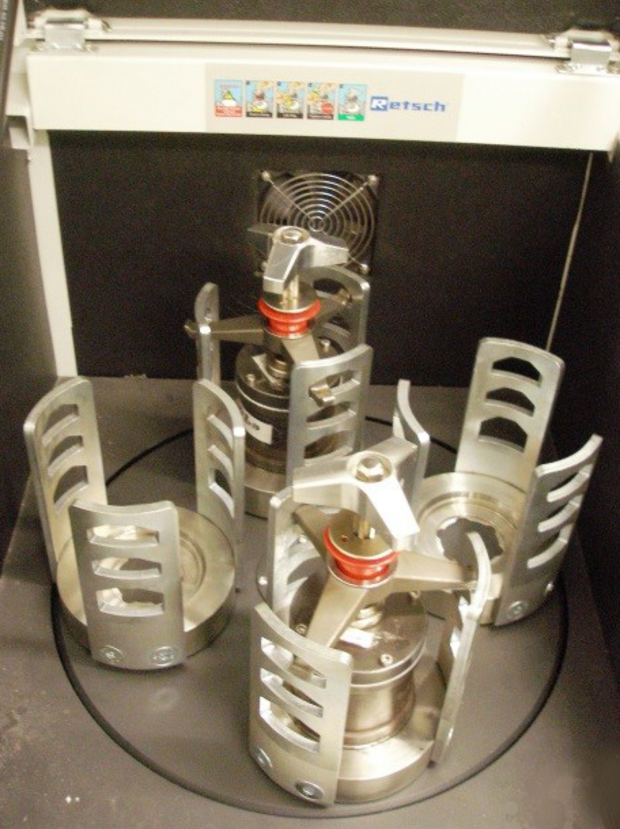 A Retsch planetary ball mill with its cover open, showing three cylindrical stainless steel grinding jars arranged on a circular platform. Each jar is wrapped in a slotted metal cage with rectangular cutouts. The components are silver-gray against a dark chamber interior.