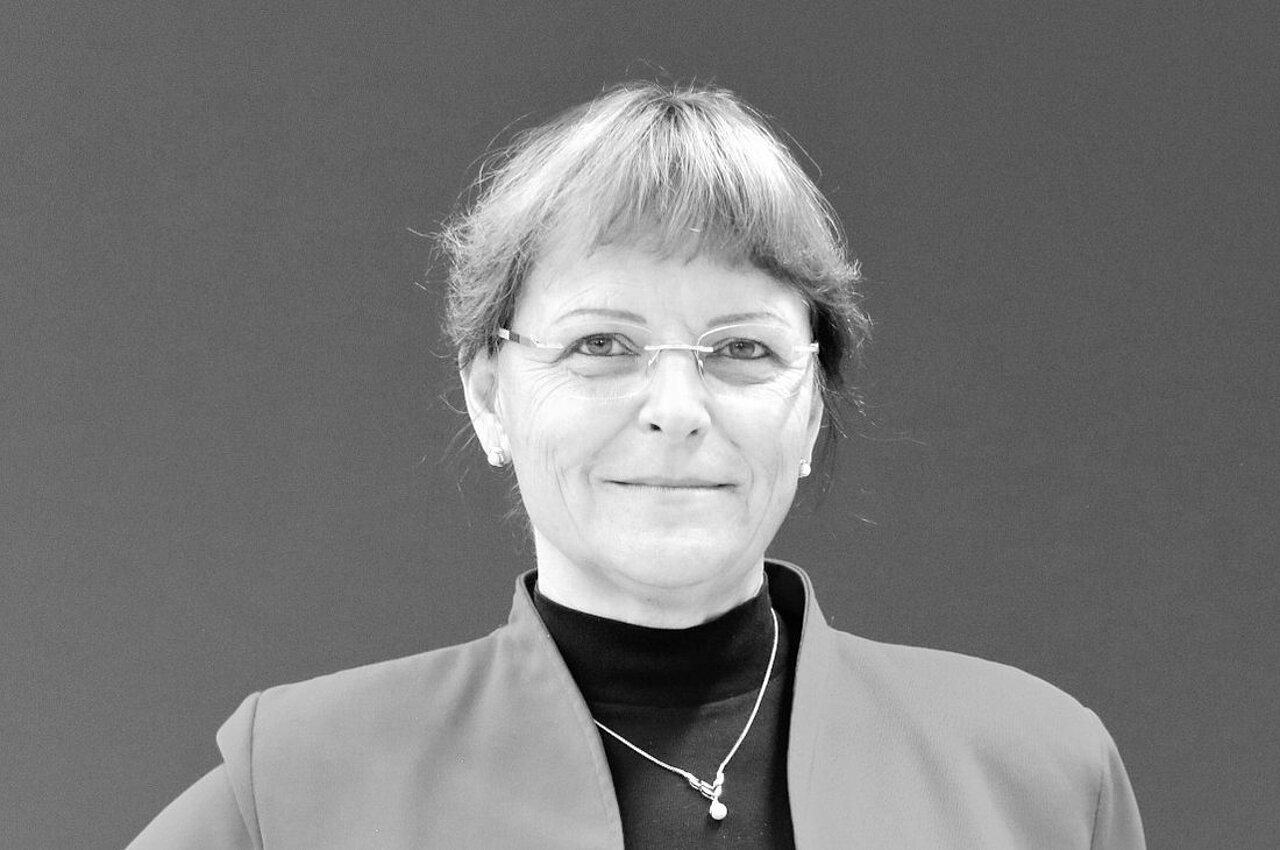 Porträt von Juliane Schmidt - Frau mit Brille, kurzem Haar und Blazer vor einfarbigem Hintergrund.