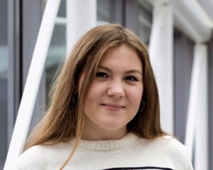 Alina Senina with long brown hair wearing a white sweater standing in front of a modern building with large windows.