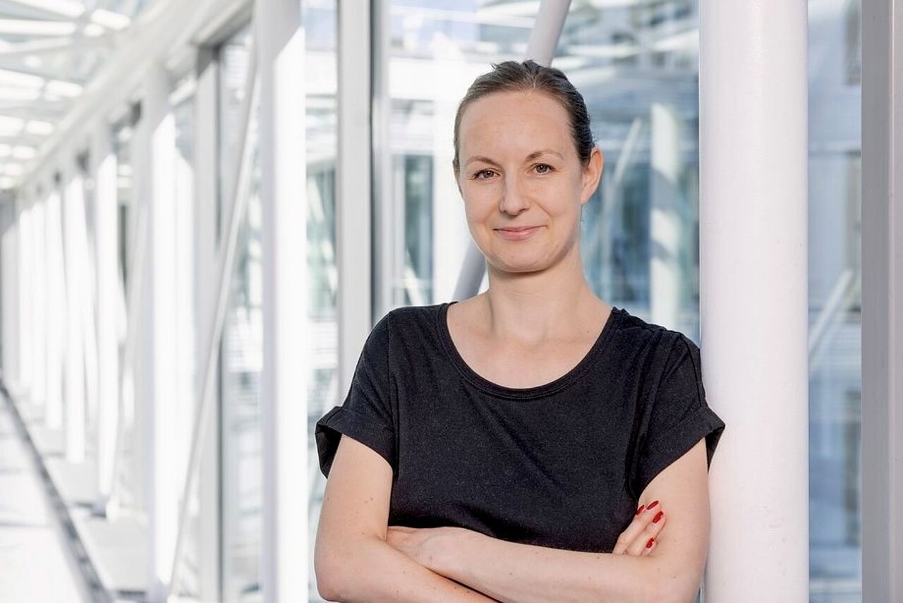 Patricia Bäuchler im IFW Dresden: eine Frau mit zusammengebundenen Haaren, die ein schwarzes T-Shirt trägt und mit verschränkten Armen in einem hellen, modernen Gebäude mit großen Fenstern steht.
